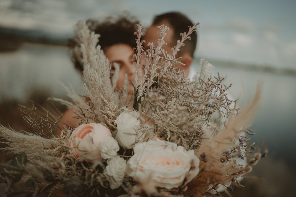 Soft-focus couple with romantic bouquet at luxury beach wedding venue, elegant floral arrangements, high-end wedding decor, dreamy coastline backdrop, romantic bridal moment.