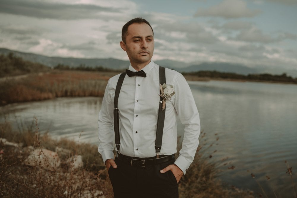 Elegant groom in classic tuxedo with suspenders and boutonnière outdoors near a serene lake, perfect for luxury wedding venues.
