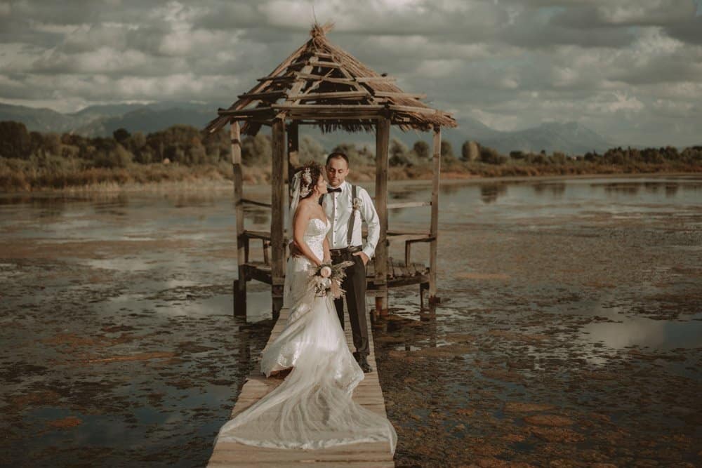 Elegant bride and groom at a luxury wedding by a serene lakeside with rustic pavilion, showcasing high-end wedding venue in nature.