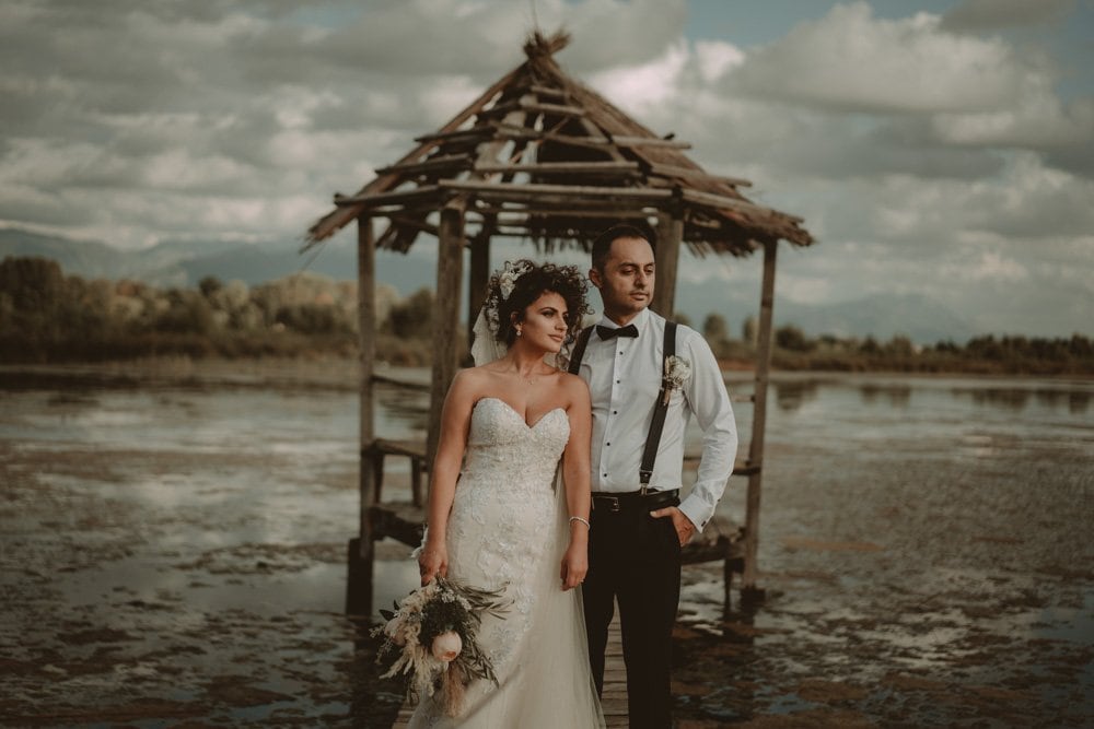 Elegant bride and groom at a luxury lakeside wedding venue, with rustic wooden gazebo and scenic water view under dramatic clouds.