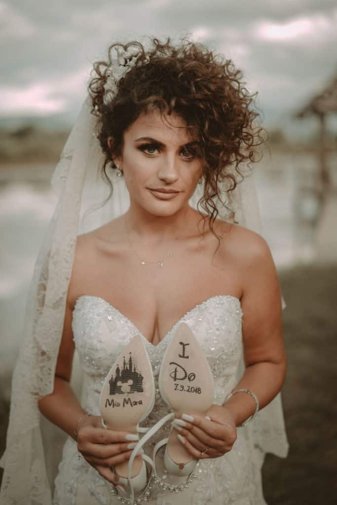 Bride holding personalized wedding shoes, outdoor beach setting, elegant wedding dress with lace details.