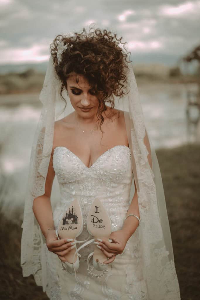 Romantic bride holding personalized wedding slippers at an outdoor seaside venue.