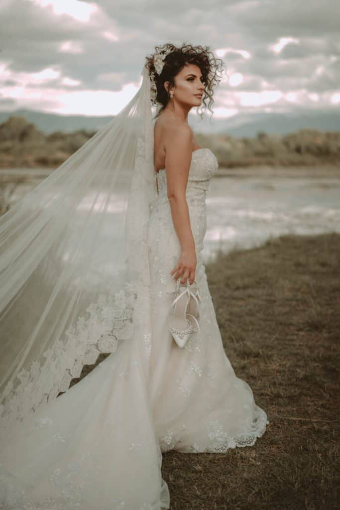 Elegant bride in a strapless lace wedding gown with a flowing veil, outdoors at sunset with scenic landscape in the background.