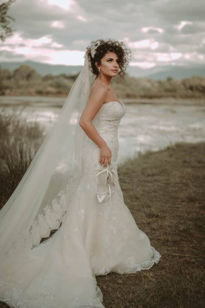 Elegant bride in a luxurious wedding gown with lace details, holding a pair of high heels outdoor at sunset. Perfect setting for high-end wedding photography and romantic bridal portraits.