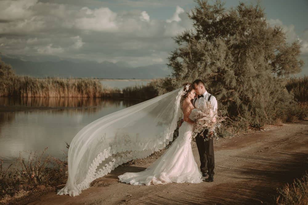 Elegant outdoor wedding photo featuring a bride and groom embracing near a serene lake with lush greenery and dramatic sky.