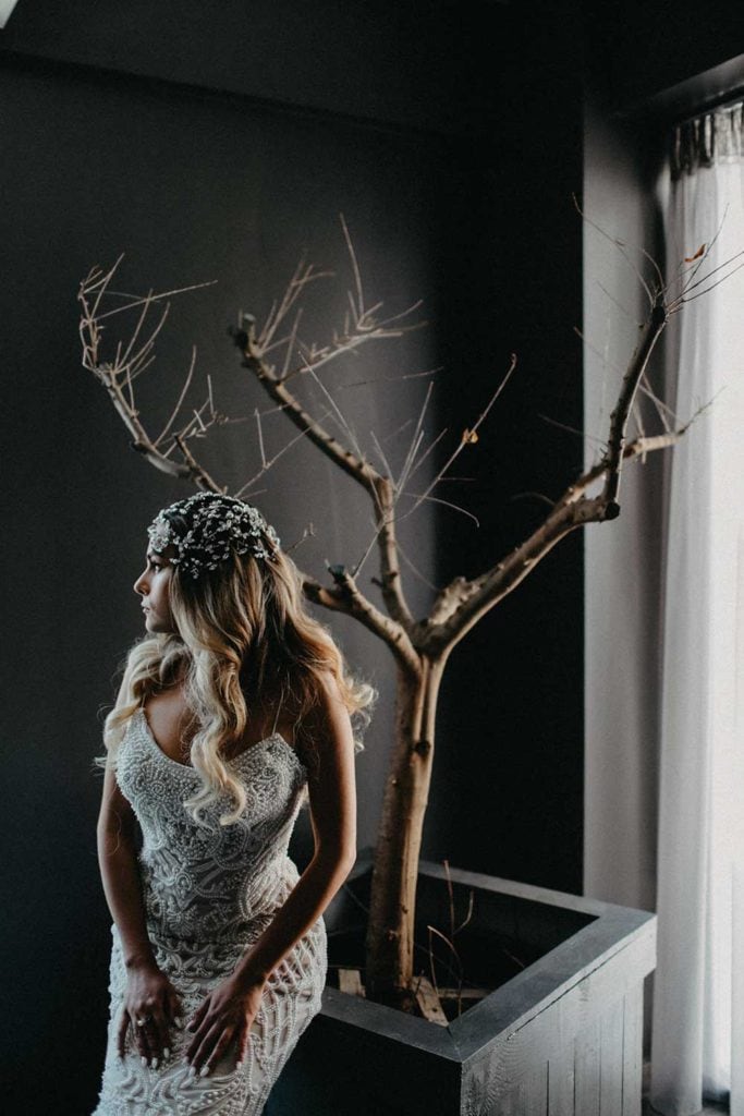 Elegant bride in intricate lace wedding gown with lush hair and floral headpiece, standing beside a minimalist indoor tree decor.