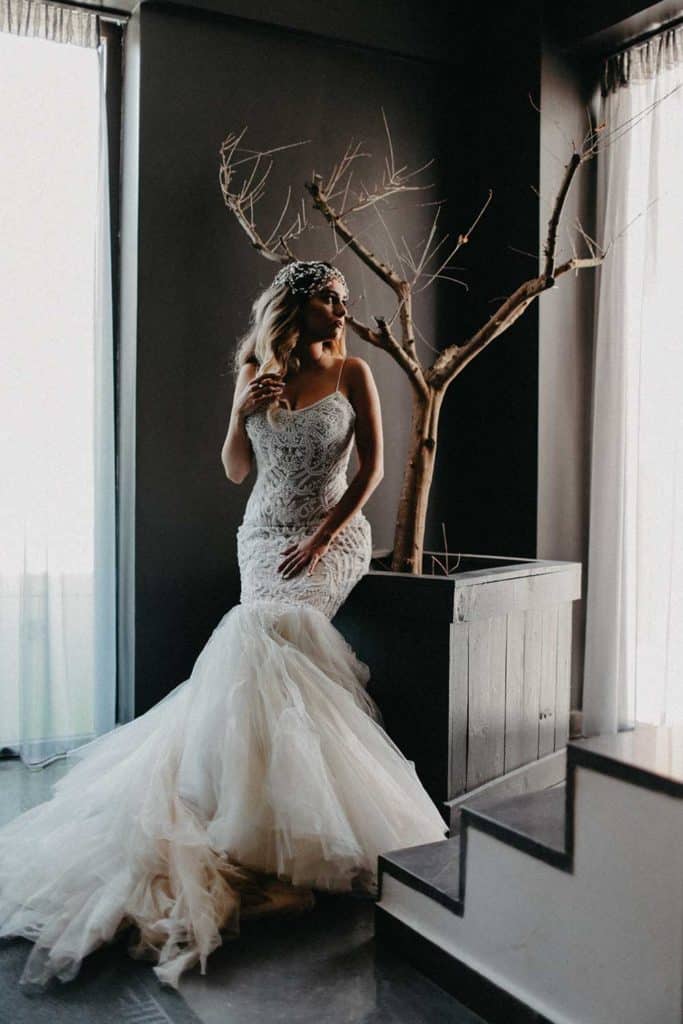 Elegant bride in a fitted lace wedding gown standing beside a modern indoor tree decor at a luxury wedding venue.