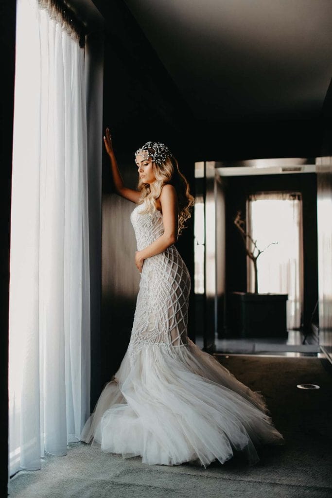 Elegant bride in a luxurious lace wedding gown standing by a sheer curtain in a sophisticated high-end venue. Perfect for a luxury wedding photoshoot.
