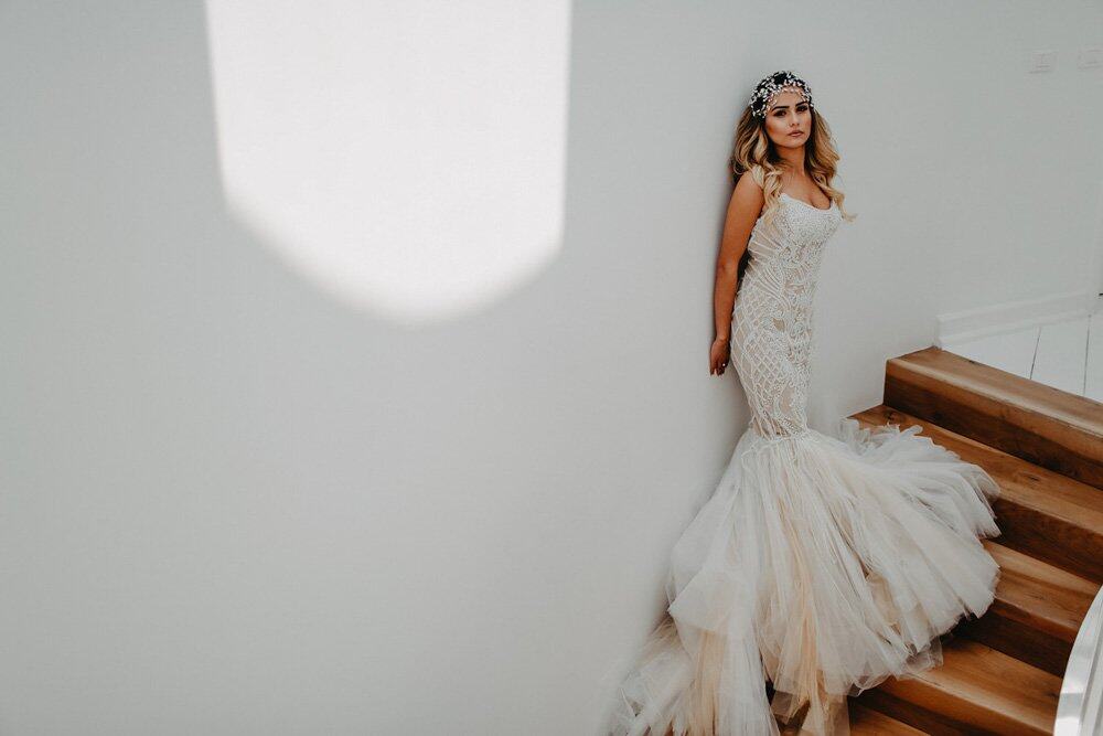 Elegant bride in a luxurious lace wedding gown standing on a staircase with modern decor.