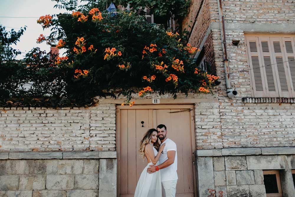 Elegant wedding couple embracing in front of a rustic stone building with vibrant orange flowers overhead.