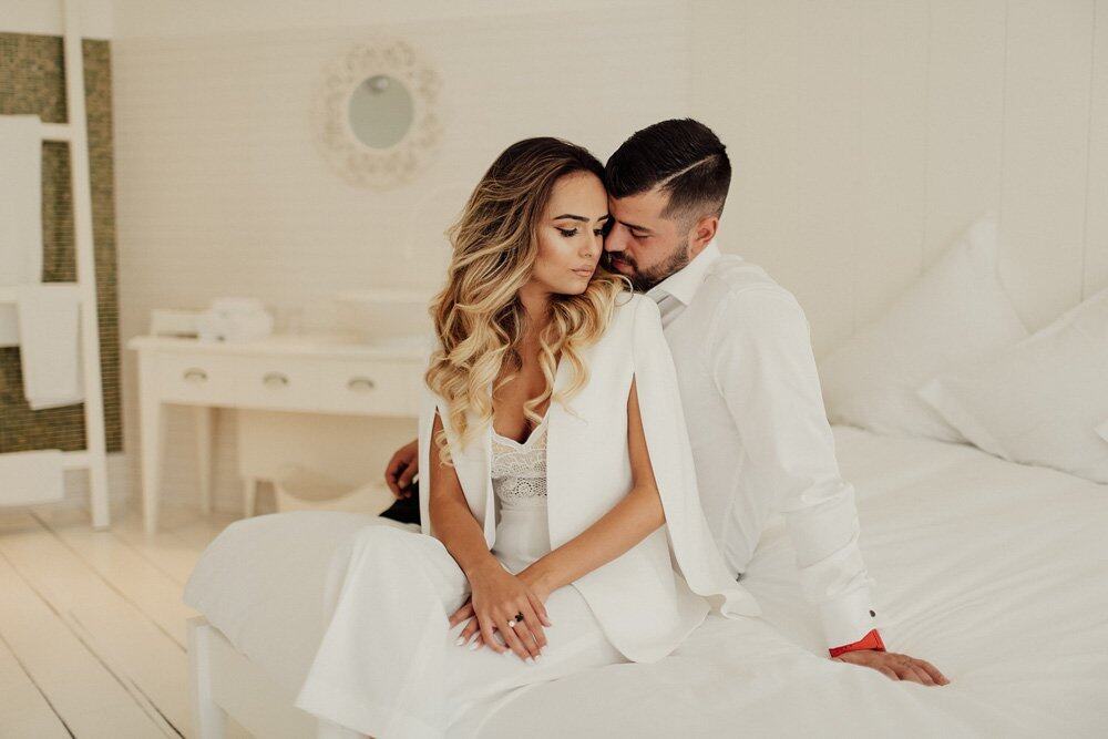 Elegant couple in white wedding attire sitting on a luxurious bed in a high-end wedding venue, surrounded by sophisticated decor.