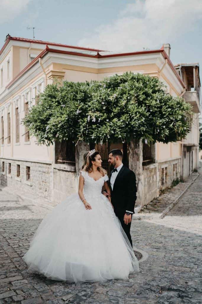Elegant wedding couple in formal attire standing beneath a lush green tree on a cobblestone street, capturing a romantic moment.