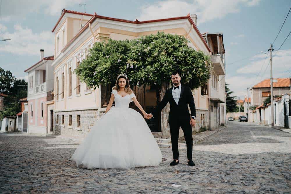 Elegant bride in a white wedding gown holding hands with groom in a black tuxedo on a cobblestone street in front of a charming historic building, capturing a luxurious wedding scene.