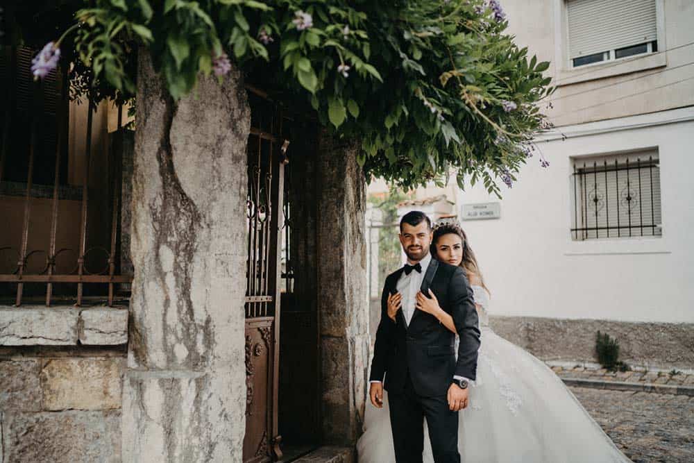 Elegant newlyweds in tuxedo and wedding gown standing outdoors under a flowering tree in front of a historic stone archway.