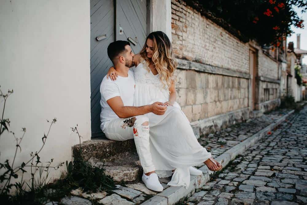 Elegant couple in white wedding attire sitting on a stone ledge in a charming, historic neighborhood with cobblestone streets.