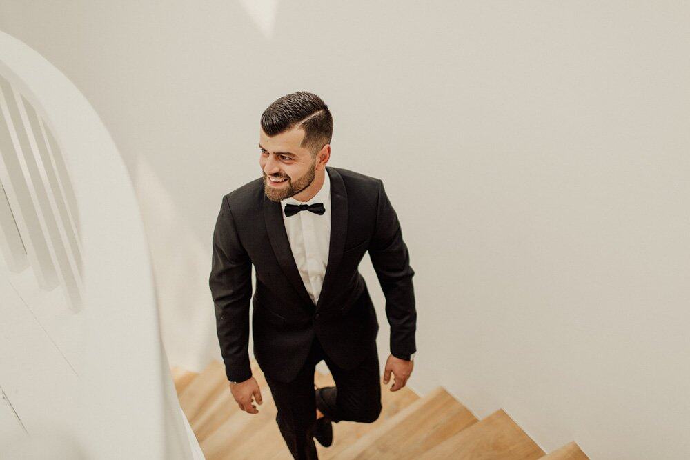 Elegant groom in classic black tuxedo ascending wooden staircase at luxury wedding venue.