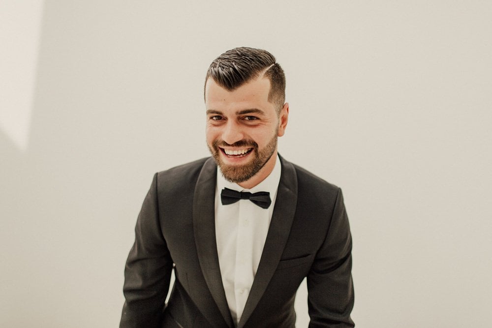 Elegant groom in a black tuxedo with a bow tie at a luxury wedding venue.