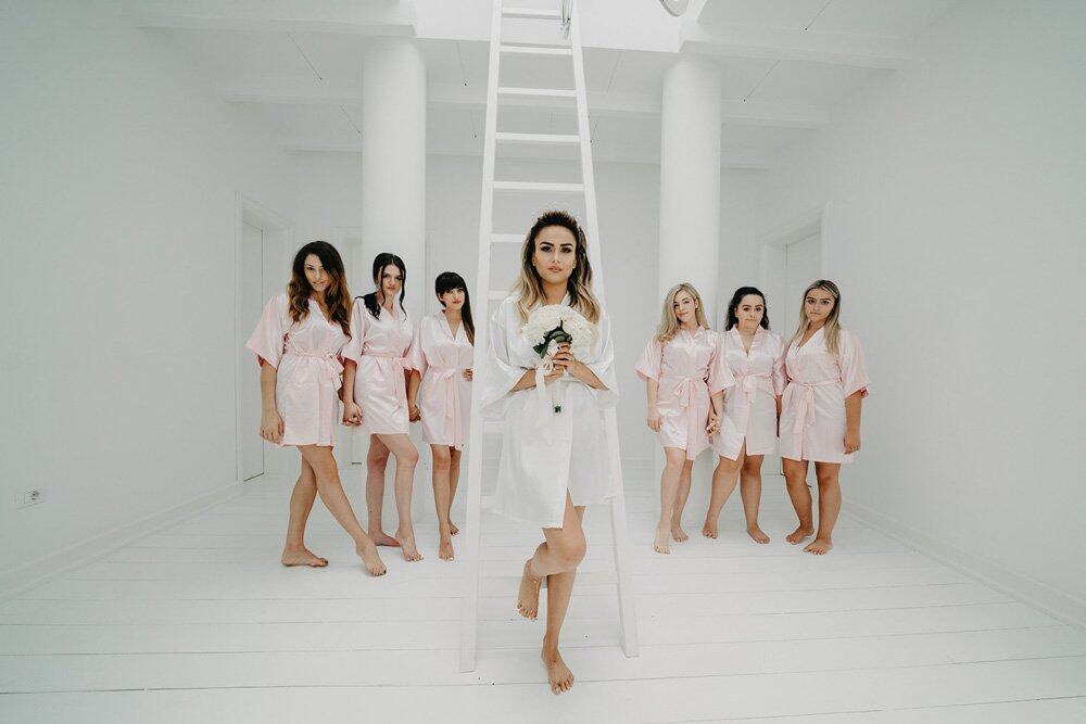 Elegant bride in white robes with bridesmaids in pastel pink, minimalist wedding prep, high-end bridal suite.