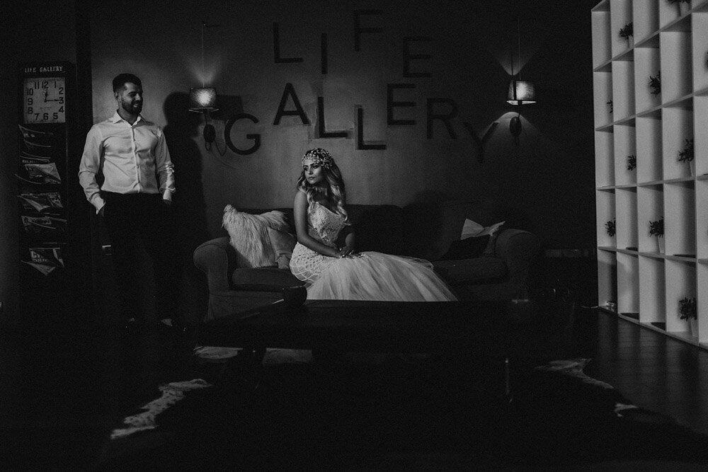 Elegant bride in a wedding gown sitting on a sofa, groom standing nearby in a stylish setting with modern decor.