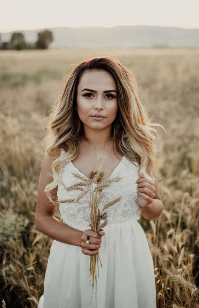 Luxury bride in a white lace dress holding dried flowers in a wheat field at sunset.