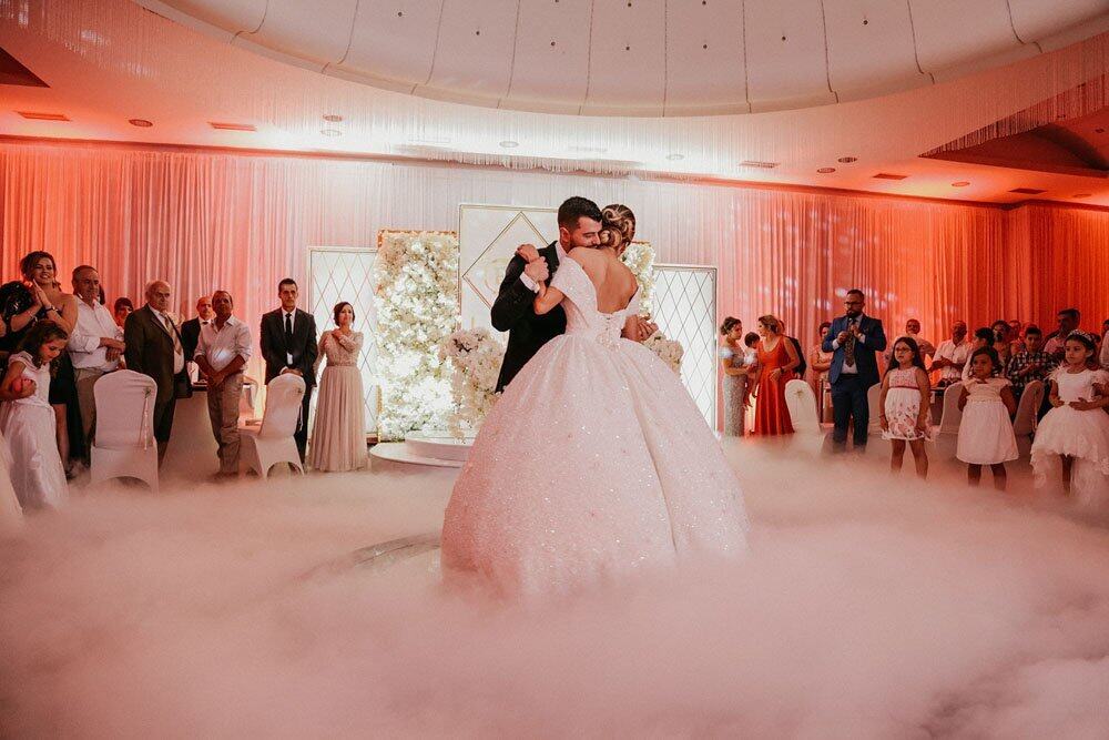 Elegant wedding couple dancing in a luxurious ballroom with dreamy fog and floral decor.