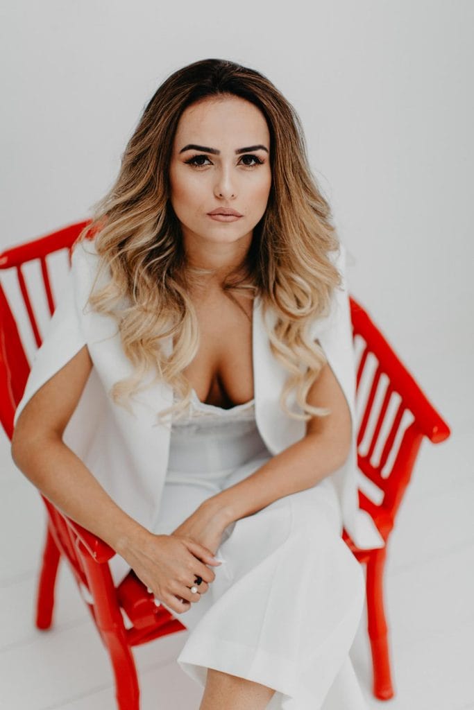 Elegant woman with styled hair and makeup, wearing a white dress, sitting on a modern red chair, perfect for luxury wedding photoshoots.