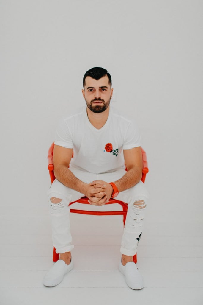 Elegant man seated on a stylish red chair, wearing a white outfit with a red floral detail, emphasizing luxury fashion and high-end wedding styling.