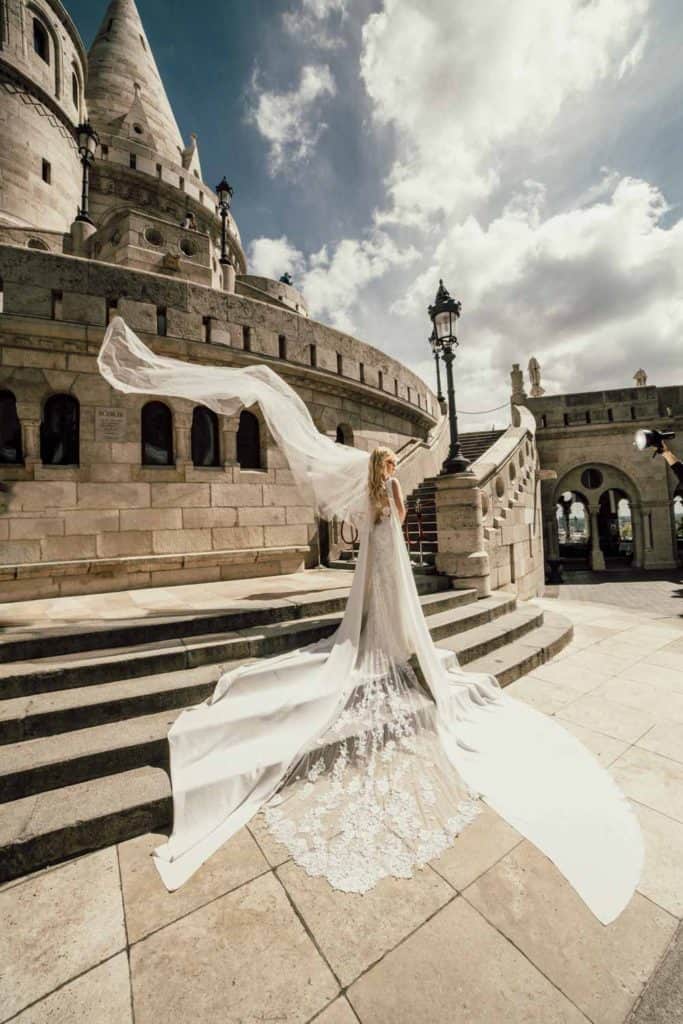 Elegant wedding gown with intricate lace train overlooking historic castle venue under a partly cloudy sky.