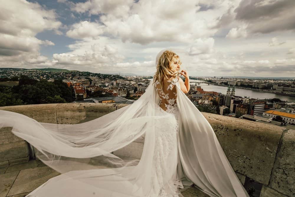 Luxury bride in an elegant wedding gown overlooking a scenic cityscape and river at a high-end venue, under dramatic cloudy skies.
