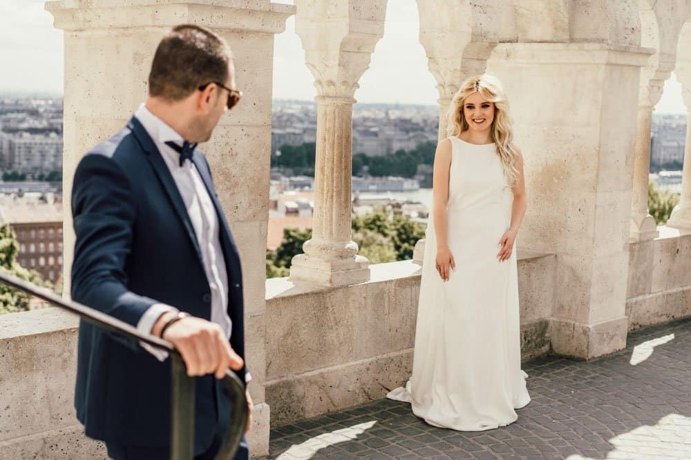 Elegant bride in white wedding gown on scenic terrace with historic architecture and city views.