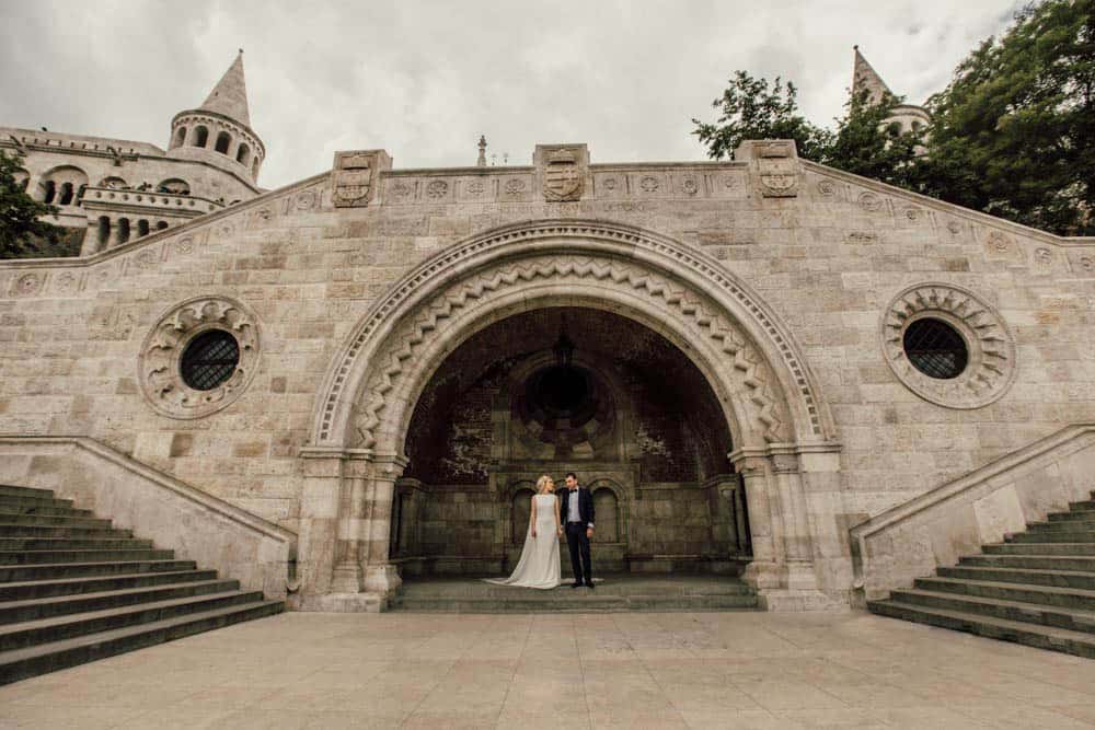 Elegant couple in wedding attire standing beneath historic castle archway, perfect luxury wedding backdrop.