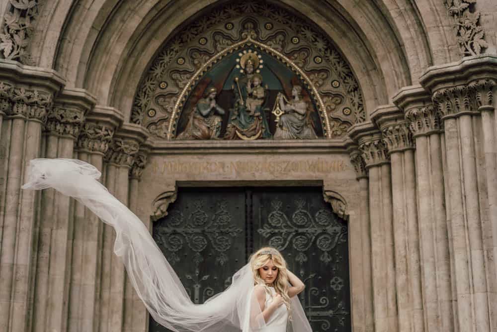 Elegant bride with flowing veil in front of historic church entrance, perfect for luxury wedding themes.