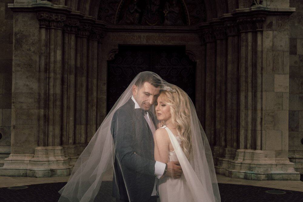 Luxurious couple in wedding attire under a veil at a historic, grand wedding venue.