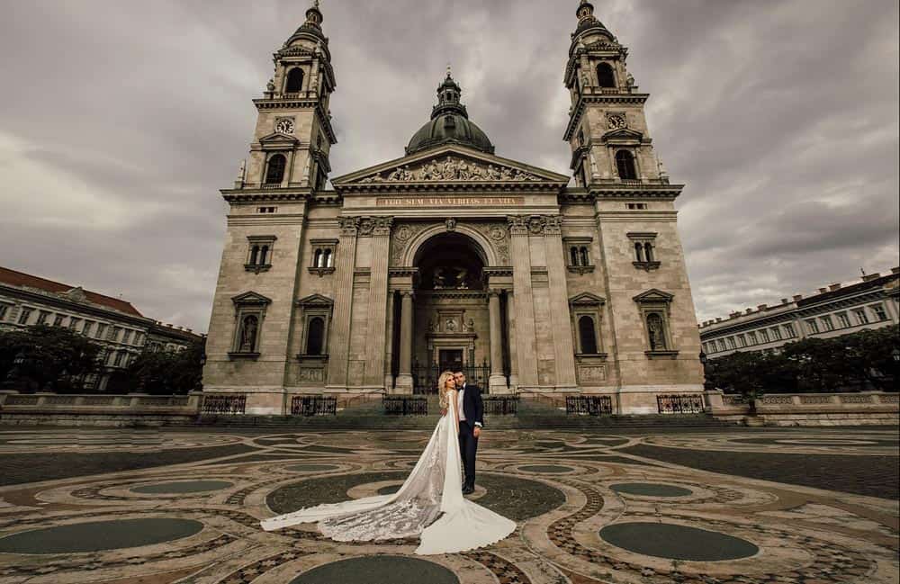Elegant wedding couple standing in front of historic basilica with dramatic cloudy sky in background.