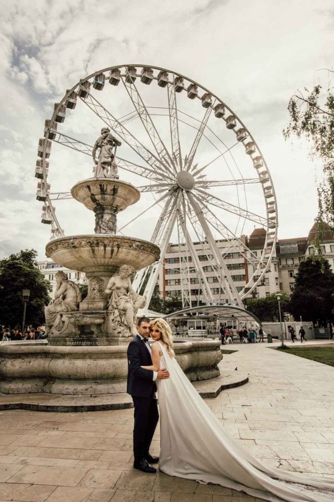 Elegant wedding couple in front of a historic fountain and modern Ferris wheel at a luxury urban park setting, perfect for upscale wedding photography.
