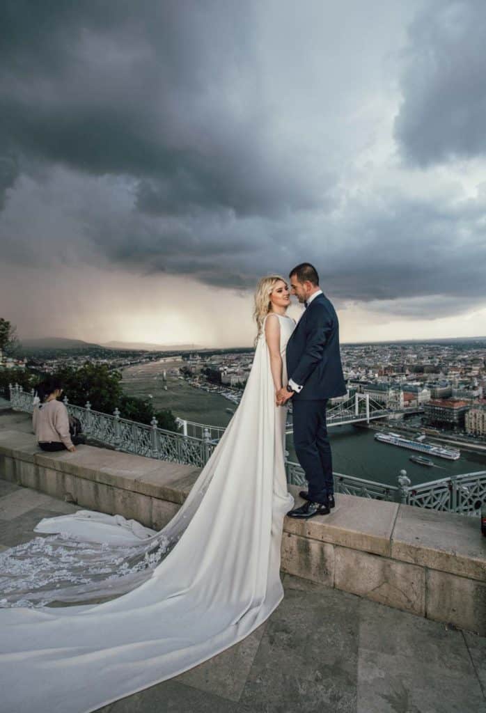 Elegant wedding couple on a high rooftop overlooking a cityscape with dramatic storm clouds, showcasing luxury wedding photography.