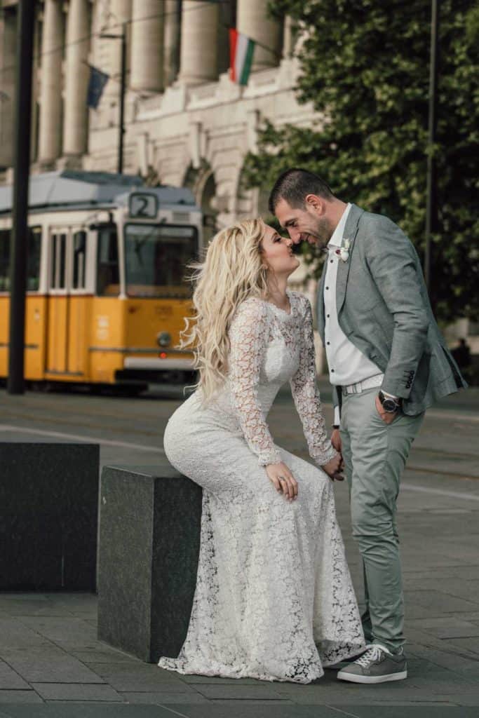 Elegant engaged couple in wedding attire sharing a moment on a city street with a tram in the background, capturing a luxurious wedding scene.