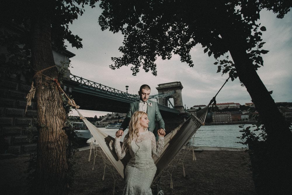 Elegant wedding couple standing beside a hammock by the river with historic bridge backdrop, capturing luxurious wedding ambiance.