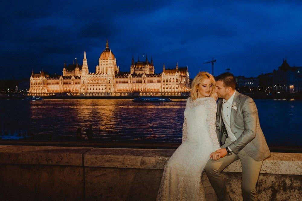 Elegant bride and groom by the Danube River with the illuminated Hungarian Parliament Building in the background, showcasing a luxurious wedding setting.