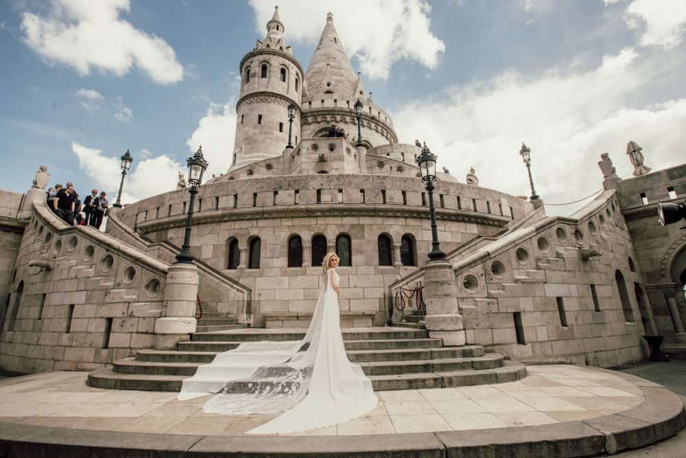 Elegant bride in a white wedding gown standing outside a fairytale castle with wedding guests in the background.