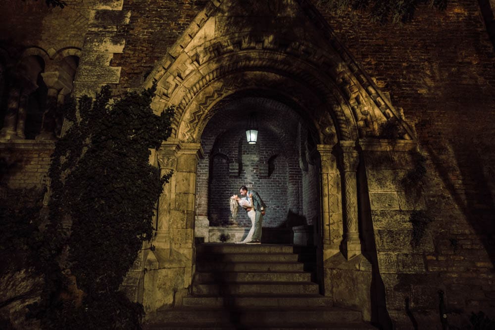 Elegant couple kissing under historic stone archway at night, perfect for luxury wedding venue photography.