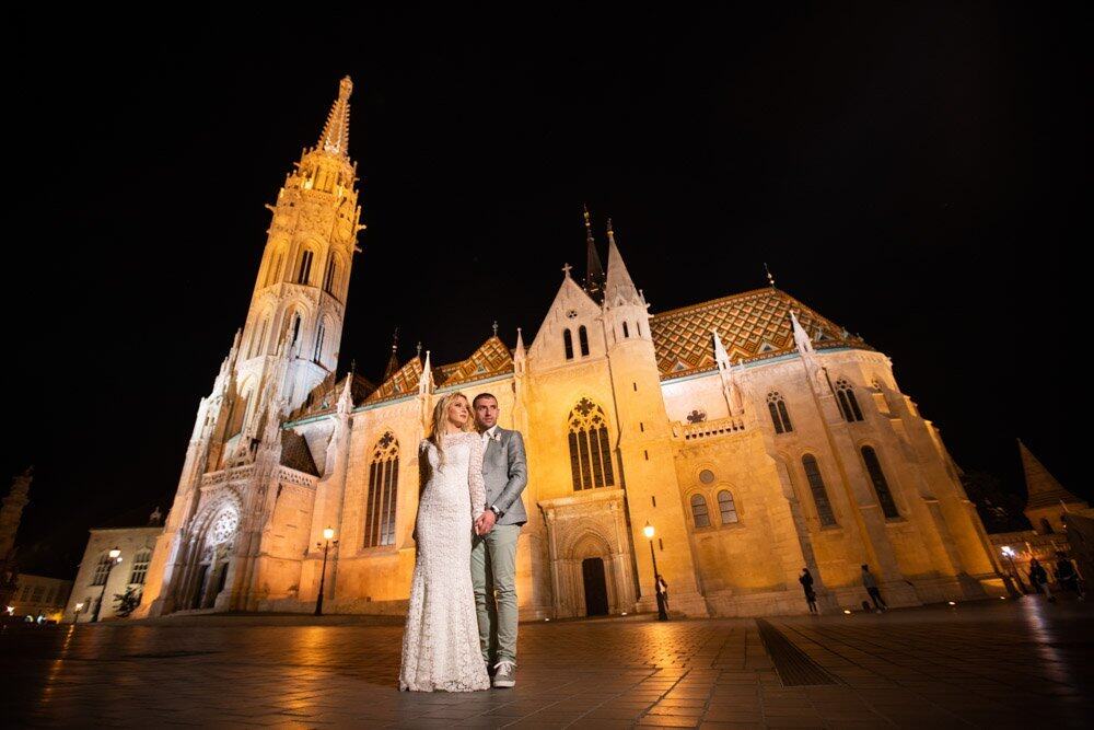 Stunning couple in elegant wedding attire outside a beautifully illuminated historic cathedral at night.