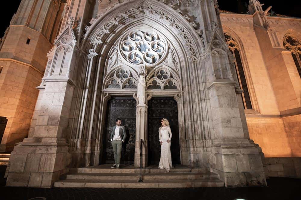 Elegant couple in wedding attire standing in front of a historic Gothic cathedral entrance at night, capturing luxury wedding moments.