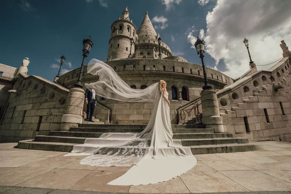 Elegant bride in a flowing white gown with a long train, standing in front of a historic castle for a luxury wedding photoshoot.