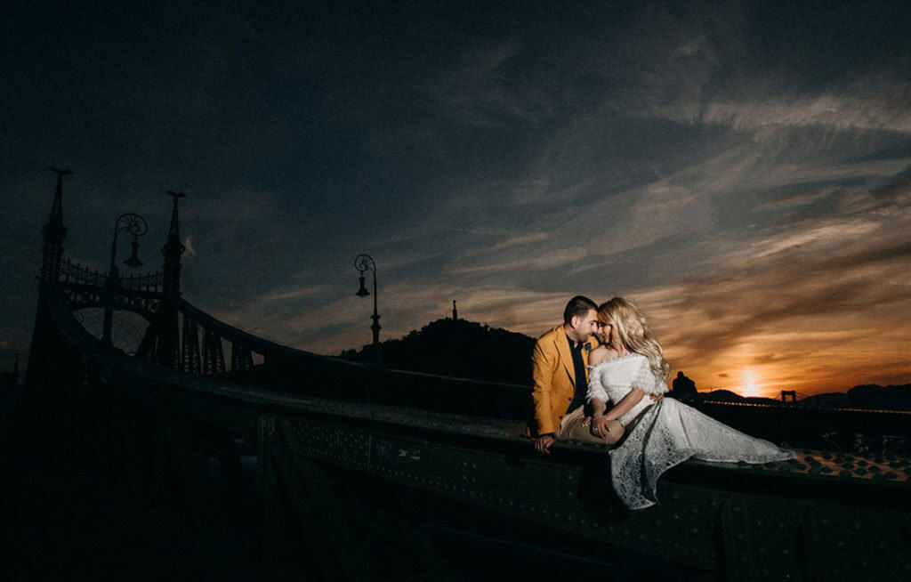 Elegant wedding couple sitting on a bridge at sunset, capturing a romantic and luxurious wedding scene.