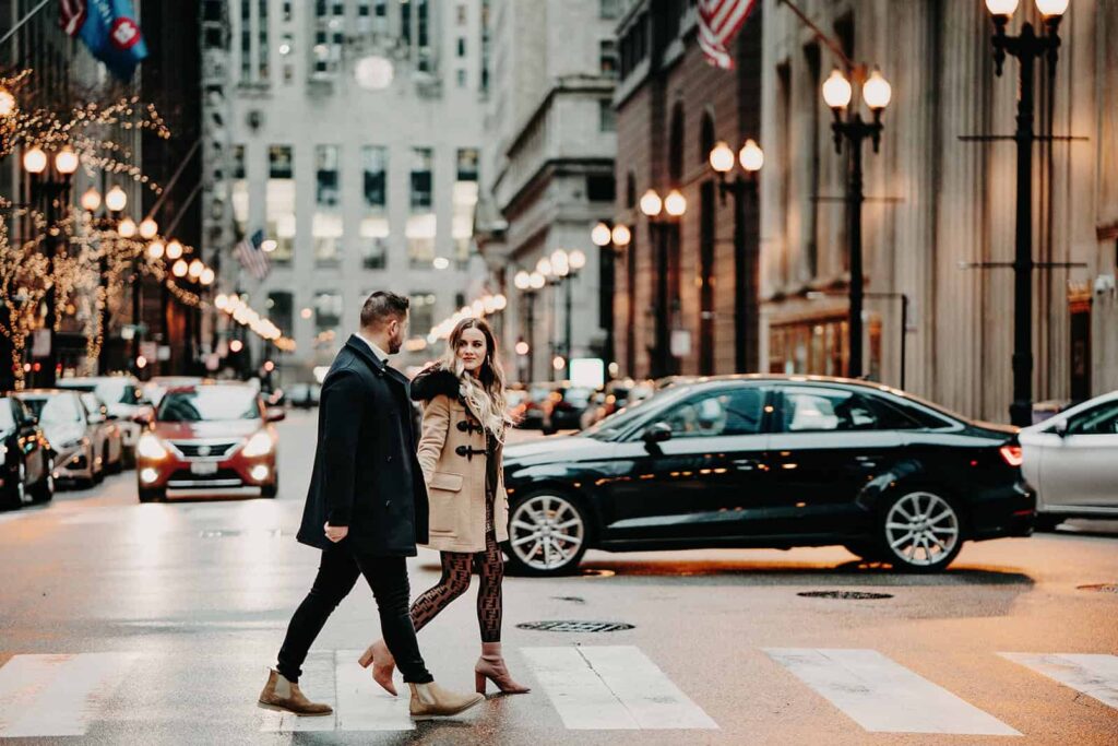Stylish couple crossing a city street at dusk with luxury cars and elegant lighting, capturing an urban romantic vibe.