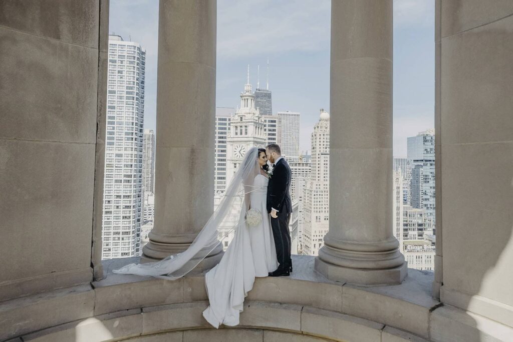 Elegant wedding couple on a rooftop with Chicago skyline view, large stone columns, luxurious high-end venue, celebrating a romantic moment.