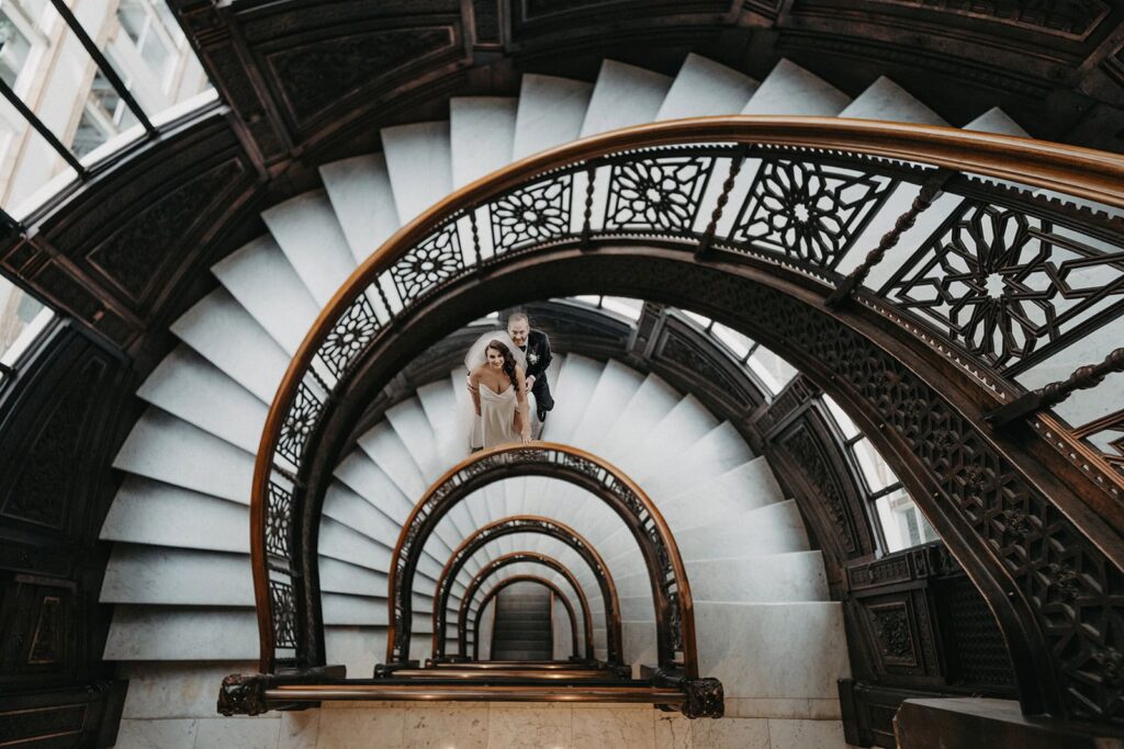 Elegant couple ascending a grand, spiraling wooden staircase in a luxury historic venue for a wedding.
