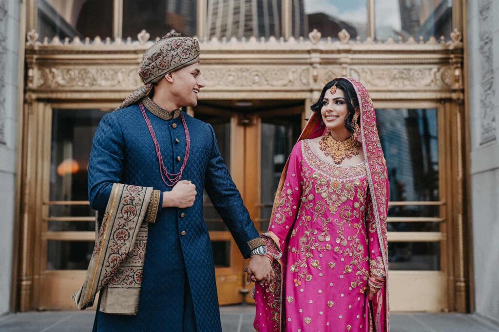 Elegant bride and groom in traditional attire holding hands outside a luxurious venue for a wedding celebration.