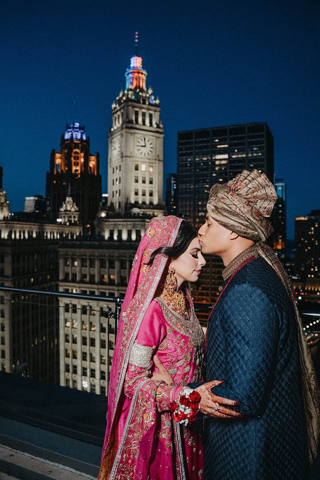 Elegant Indian bride and groom in traditional attire sharing a romantic moment on rooftop with city skyline at night.
